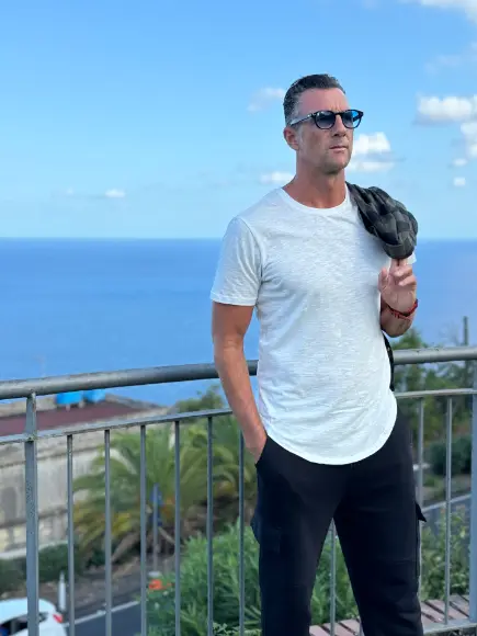 Mann mit weißem T-Shirt und schwarzer Hose, trägt Sonnebrille und Jacke über der Schulter